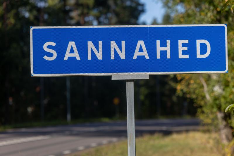 En blå skylt med vita bokstäver visar namnet ”Sannahed” vid en väg med träd i bakgrunden.