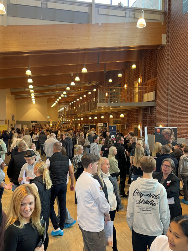 Folkmassa som deltar i ett inomhusevenemang i en stor, väl upplyst hall med högt i tak och trädetaljer. Vissa tittar på montrar och utställningar, medan andra går omkring eller pratar.