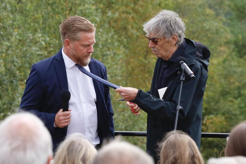 En äldre man granskar slipsen på en yngre man som håller i en mikrofon under ett utomhusevenemang med publik.