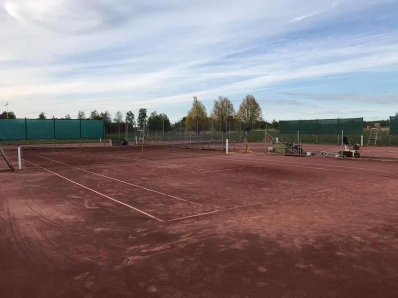 En tennisbana med grus utomhus med lite utrustning och verktyg utspridda runt omkring, omgiven av gröna staket och träd i bakgrunden under en delvis molnig himmel.