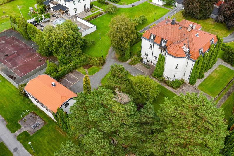 Flygfoto över ett stort vitt hus med rött tak, omgivet av träd och grönska, intill ett uthus och en inhägnad tennisbana.