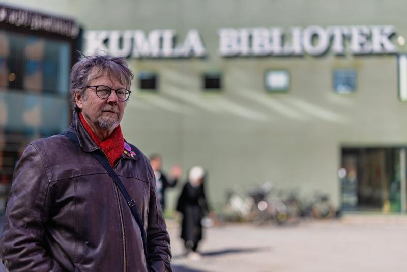 En man i brun skinnjacka och röd halsduk står utanför Kumla Bibliotek, med cyklar och människor suddiga i bakgrunden.