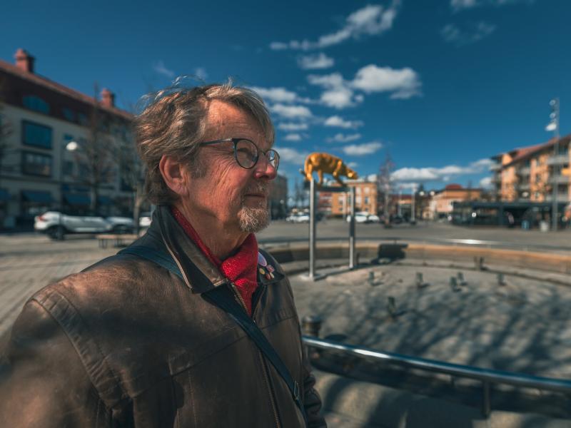 En äldre man med glasögon och brun skinnjacka står på ett torg i en stad med en gul djurskulptur i bakgrunden.