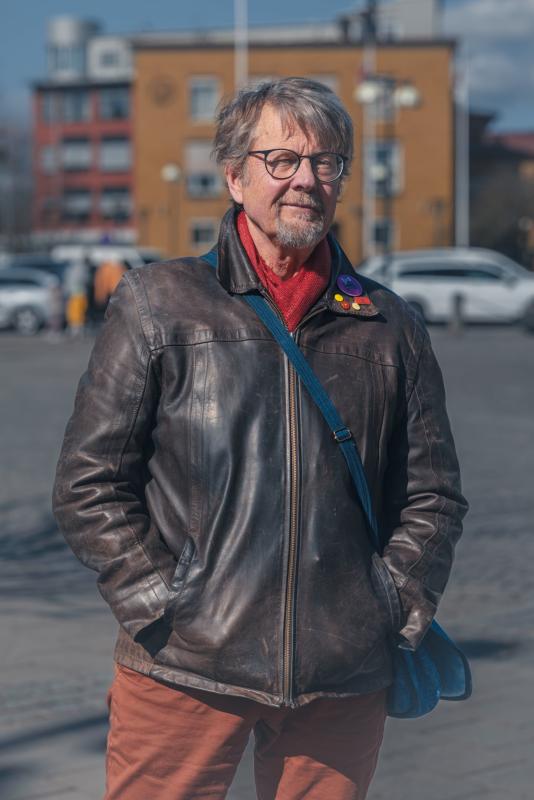En äldre man med glasögon, skinnjacka och en blå axelremsväska står utomhus en solig dag med byggnader och parkerade bilar i bakgrunden.