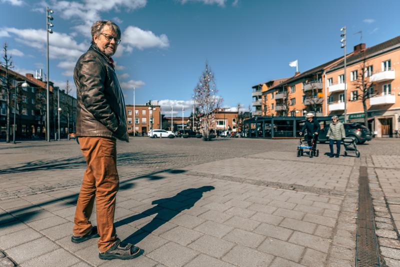 En man i skinnjacka och orange byxor står på ett asfalterat torg och tittar bakåt. I bakgrunden går två personer med barnvagn i närheten av parkerade bilar och byggnader.