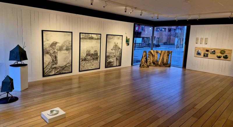 Konstgalleriinteriör med inramade skisser på en vit vägg, skulpturer på montrar och ett trägolv. Utesikt genom ett stort fönster i bakgrunden.