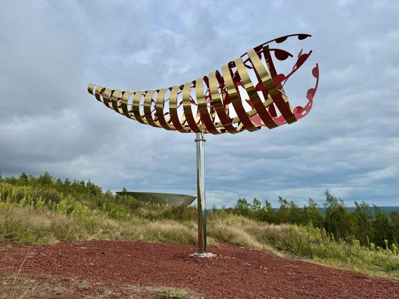 En stor, abstrakt metallskulptur som liknar en ribbad, böjd form står på en stolpe i ett gräsbevuxet utomhuslandskap under en molnig himmel.