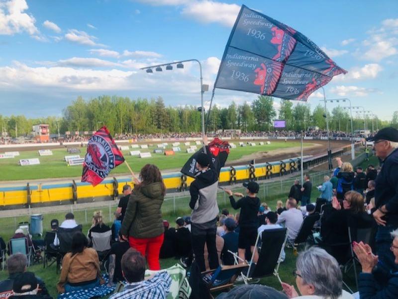 Åskådare tittar på ett motorsportevenemang på en utomhusbana, med några människor som viftar med Indiana Speedway-flaggor på läktarna under en delvis molnig himmel.