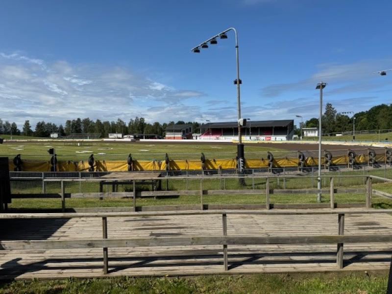 Speedwaystadion utomhus med en tom bana, träläktare i förgrunden, barriärer och säkerhetsstängsel samt byggnader i bakgrunden under en delvis molnig himmel.