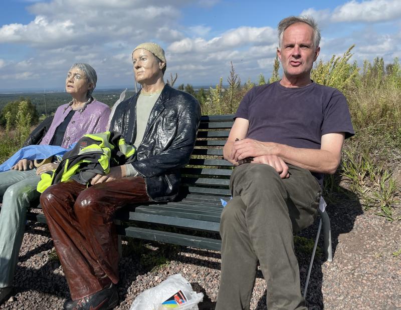 En man sitter på en bänk utomhus bredvid två verklighetstrogna statyer av människor, med buskar och en blå himmel i bakgrunden.