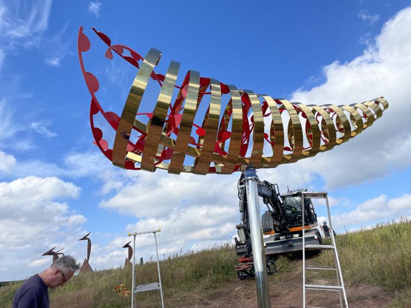 En man står nära en stor, abstrakt metallskulptur som håller på att installeras på ett öppet fält, med en blå himmel och spridda moln i bakgrunden.