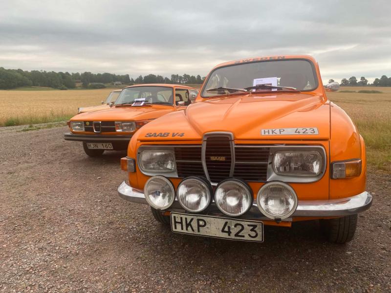 Två orange Saab-bilar i retrostil står parkerade på en grusväg nära en åker. Den främre bilen har fyra rallystrålkastare och båda bilarna har registreringsskyltar.
