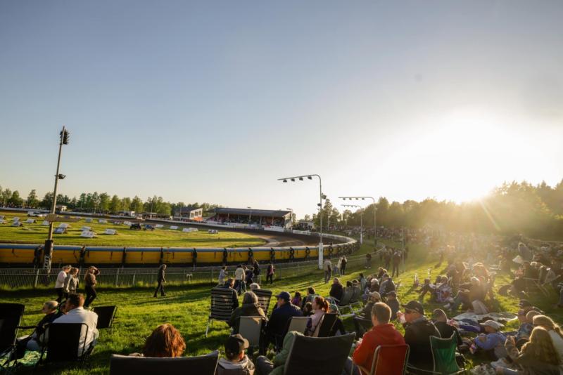 Åskådare sitter på en gräsbevuxen kulle och tittar på en motorsporttävling på en utomhusbana under sen eftermiddag med solen lågt på himlen.