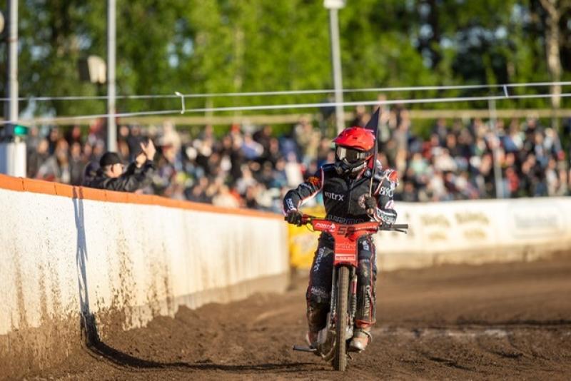 En motorcykelförare i full utrustning kör på en grusbana med en mängd åskådare i bakgrunden.