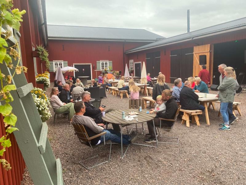 Människor i olika åldrar sitter vid runda träbord på en innergård mellan röda byggnader och pratar och kopplar av en molnig dag. I förgrunden syns några blommor och grönska.