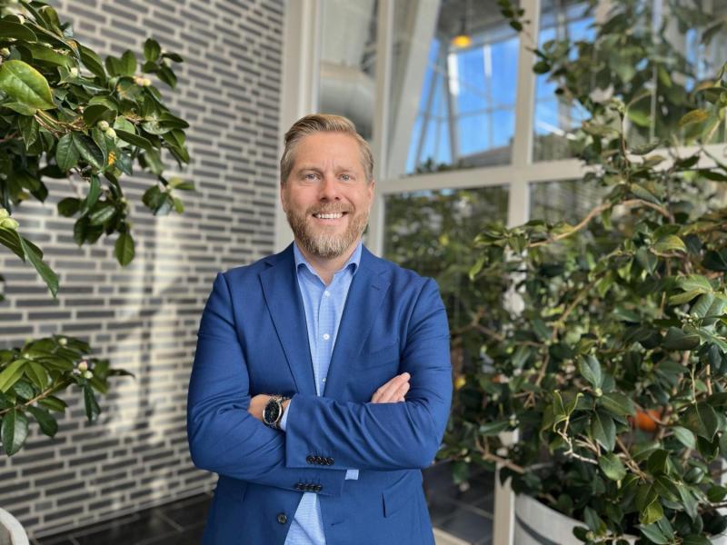 En man i blå kostym står med armarna i kors och ler i en inomhuslokal med stora fönster och gröna växter.