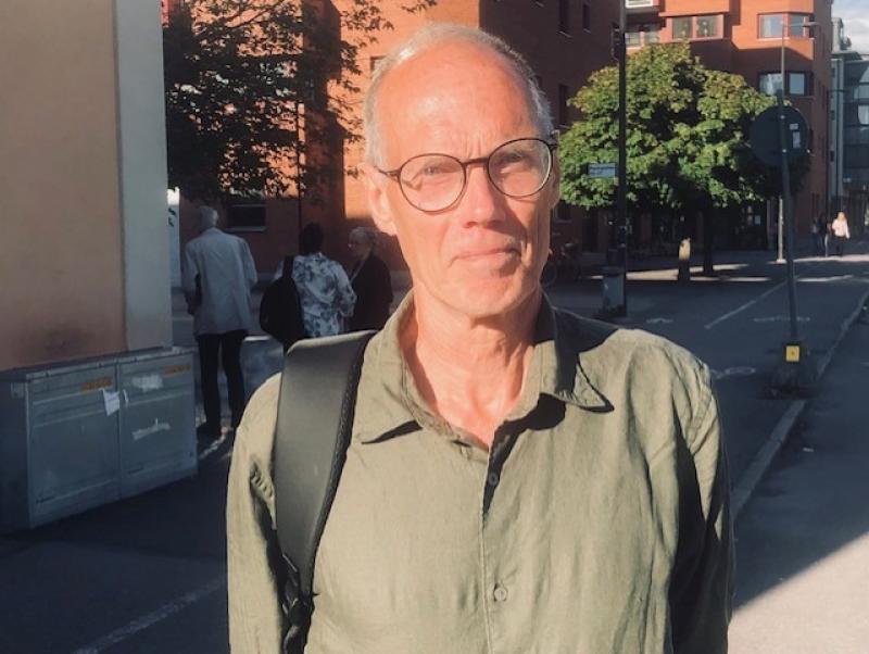 En man med glasögon och grön skjorta står utomhus på en solig gata med byggnader och träd i bakgrunden.