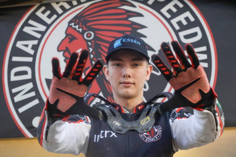 En ung speedwayförare i tävlingskläder står med en nackkrage i handen framför en skylt med texten ”Indianerna Speedway” och en logotyp föreställande en indianhövding.