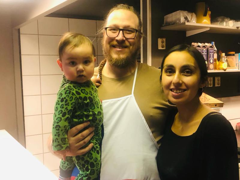En man som håller ett barn står bredvid en kvinna i ett kök, med hyllor med matlagningsingredienser och redskap i bakgrunden.