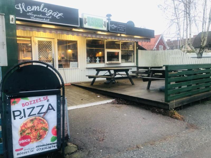 Utomhusvy av en liten restaurang med en grön uteplats, picknickbord och en pizzareklam. Byggnaden har en rustik exteriör med flera skyltar och ljus synliga.