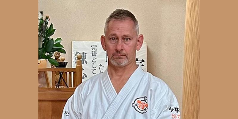 Medelålders man i vit kampsportsuniform står inomhus, med en liten helgedom och japansk kalligrafi i bakgrunden.