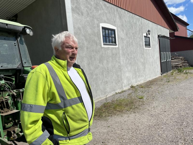 En äldre man i en gul varseljacka står utanför bredvid en byggnad och en grön traktor en solig dag.