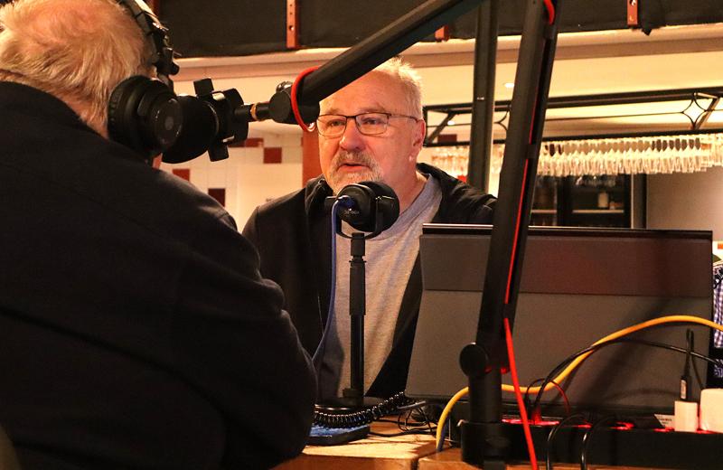 Två män med hörlurar sitter vid ett bord med mikrofoner och inspelningsutrustning och ser ut att föra ett samtal eller spela in en podcast i en inomhusmiljö.