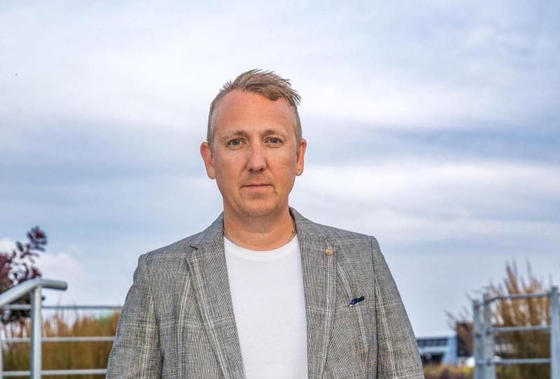 En man klädd i en grå rutig kavaj och en vit skjorta står utomhus framför en molnig himmel och några växter.