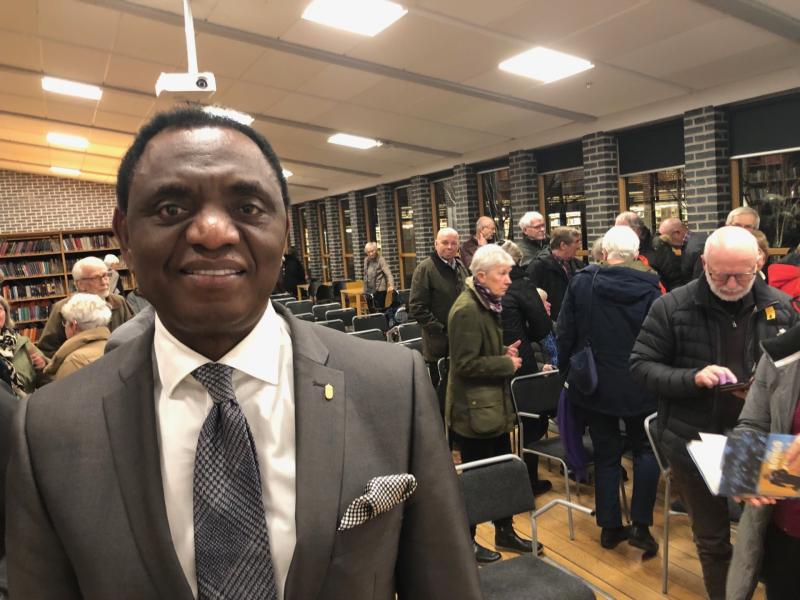 En man i kostym står i förgrunden av ett rum fyllt med människor som minglar.