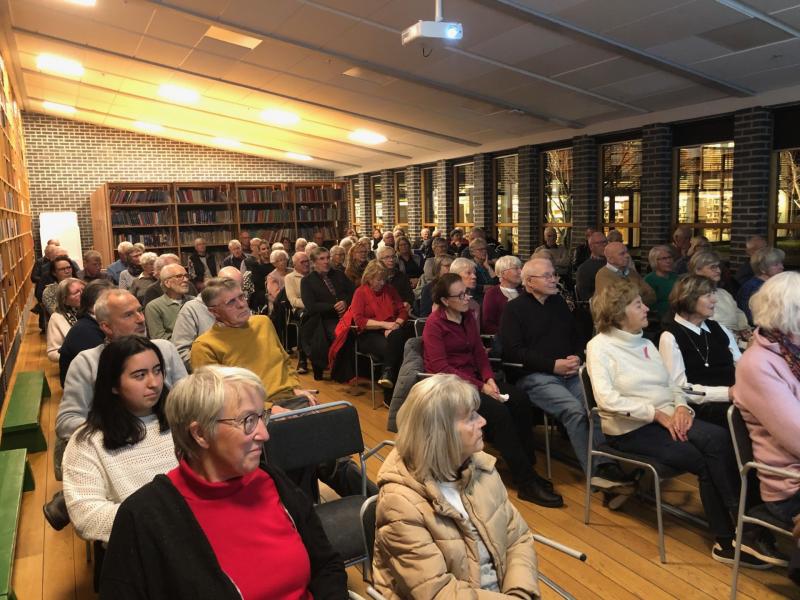 En grupp människor sitter uppmärksamt i ett bibliotek, vända mot en projektorduk. Bokhyllor kantar väggarna och stora fönster syns i bakgrunden.