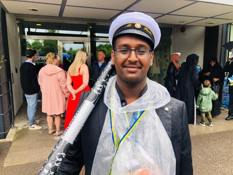En ung man i examensmössa och klar regnponcho står och ler framför en byggnad, omgiven av människor. Han bär glasögon, en medalj och håller en upprullad affisch.