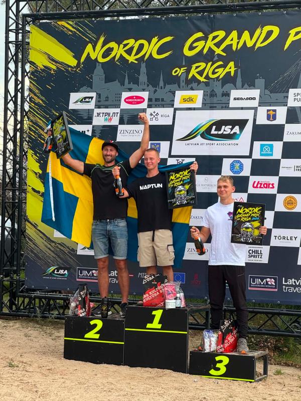Tre män står på podier med pokaler och en svensk flagga i händerna framför en bakgrund med motiv från Nordiska Grand Prix-tävlingen och firar sina placeringar i tävlingen.