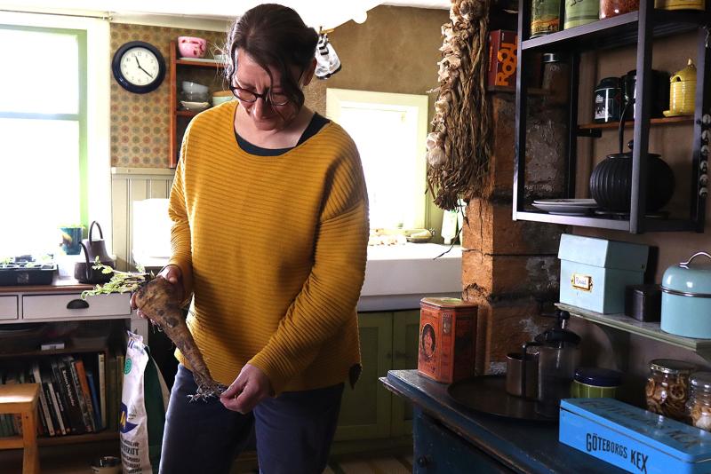 En kvinna i gul tröja inspekterar en stor rotfrukt i ett rustikt kök med hyllor, diskbänk och olika köksredskap.