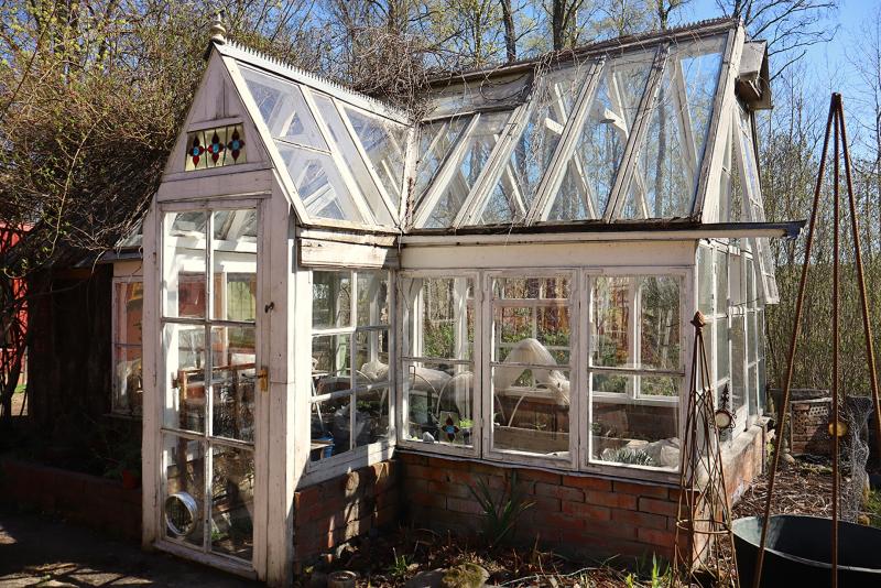 Ett glasväxthus med tegelbas och triangulära takpaneler står i solljus, omgivet av växter och träd.