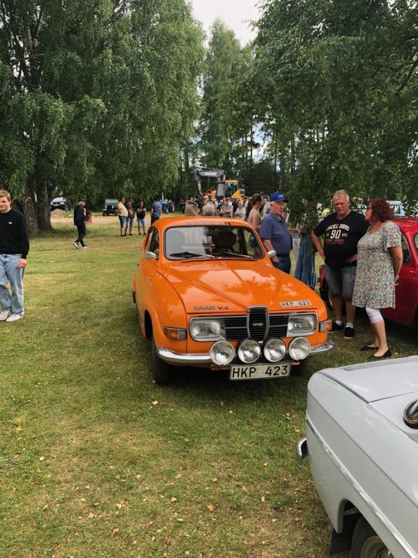 En orange Saab V4 står parkerad på gräset bland träden, omgiven av människor vid vad som verkar vara en bilutställning eller sammankomst utomhus.