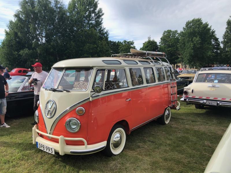 En vintage-modell av en röd och vit Volkswagen-husbil med takräcke står parkerad på gräset bland andra klassiska bilar, och det står människor i närheten.