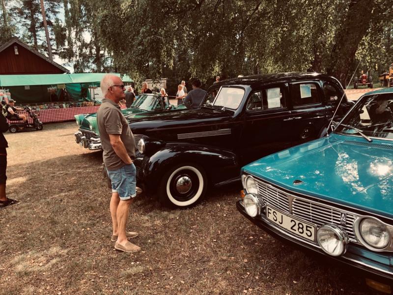 En man står utomhus och betraktar en rad klassiska bilar som står parkerade på gräset, med träd och en träbyggnad i bakgrunden.