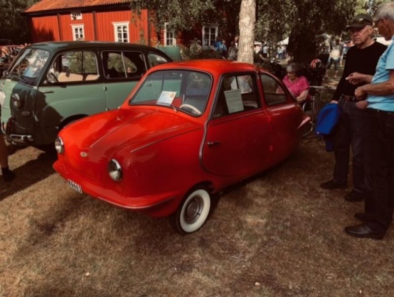 En liten, vintage-röd trehjulig bil står parkerad på gräset bredvid en grön bil, och i närheten står människor vid ett utomhusarrangemang.