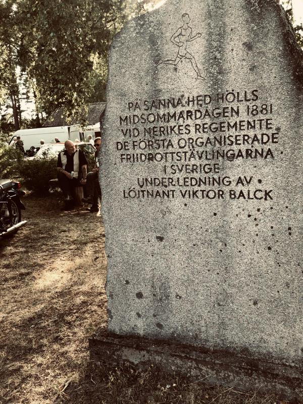 Ett stenmonument med en svensk inskription till minne av den första organiserade friidrottstävlingen i Sverige år 1881, med människor och träd synliga i bakgrunden.