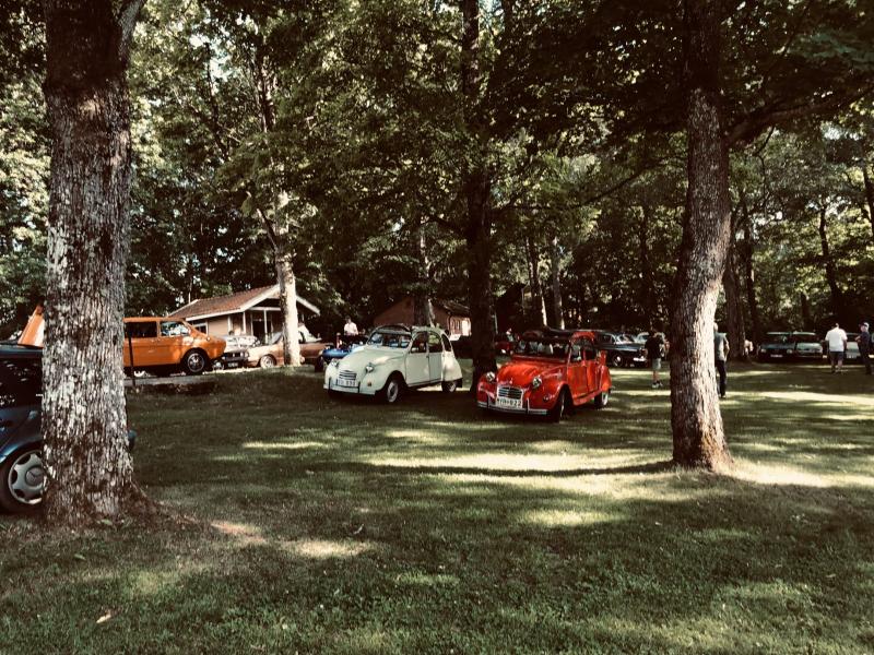 Klassiska bilar, däribland en vit och en röd Citroën 2CV, står parkerade på en gräsplätt under träden nära ett hus, med människor i bakgrunden.