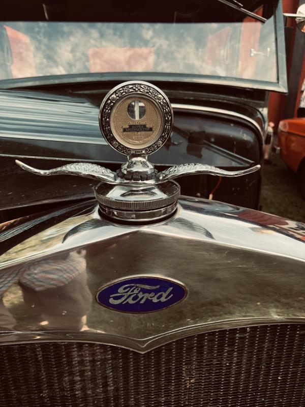 Närbild på en klassisk kylgrille från en Ford-bil, med en blå Ford-logotyp och en utsmyckad motorhuvsdekoration med en temperaturmätare.