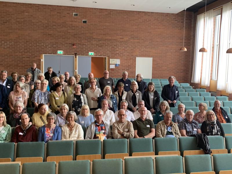 En stor grupp vuxna poserar för ett foto i en aula med gröna stolar, tegelväggar och höga fönster.