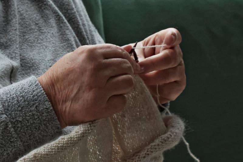 Närbild av en äldre person som stickar med vitt garn, klädd i en grå tröja, mot en grön bakgrund.