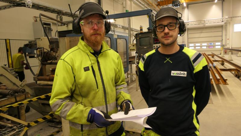 Två män i skyddsutrustning och arbetsuniform står inomhus i en industrianläggning och håller i dokument, med maskiner och metallbalkar i bakgrunden.