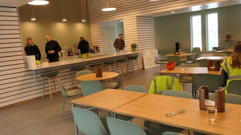 Flera personer står vid en disk i en modern kafeteria med träbord, gröna stolar och minimalistisk inredning. Några jackor med hög synlighet är draperade över stolarna.