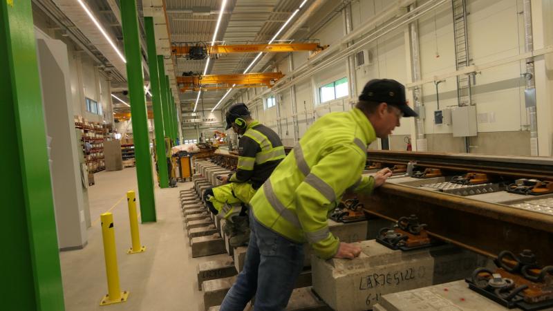 Två arbetare i varseljackor och hjälmar inspekterar och arbetar på en del av ett järnvägsspår inomhus, med industriutrustning synlig i bakgrunden.