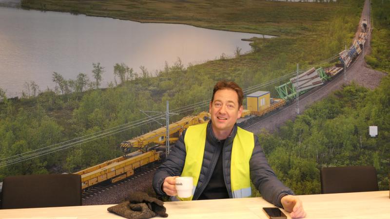 En man i gul skyddsväst sitter vid ett bord med en kaffekopp i handen, en telefon och handskar framför sig. På väggen bakom honom finns en väggmålning av ett godståg i ett lantligt landskap.