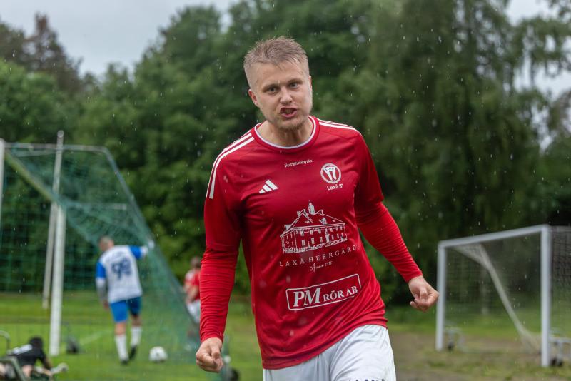 En fotbollsspelare i en röd tröja verkar ropa eller jubla på en regnig plan, med målstolpar och en annan spelare i bakgrunden.