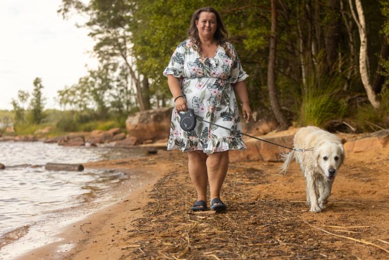 En kvinna i en blommig klänning promenerar med en golden retriever längs en sandstrand vid en sjö, omgiven av träd.