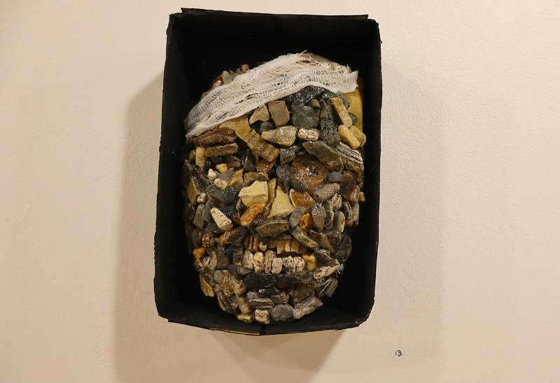 Ett skulpterat ansikte gjort av olika små stenar är placerat i en svart rektangulär låda med en bit vit gasbinda i pannan.